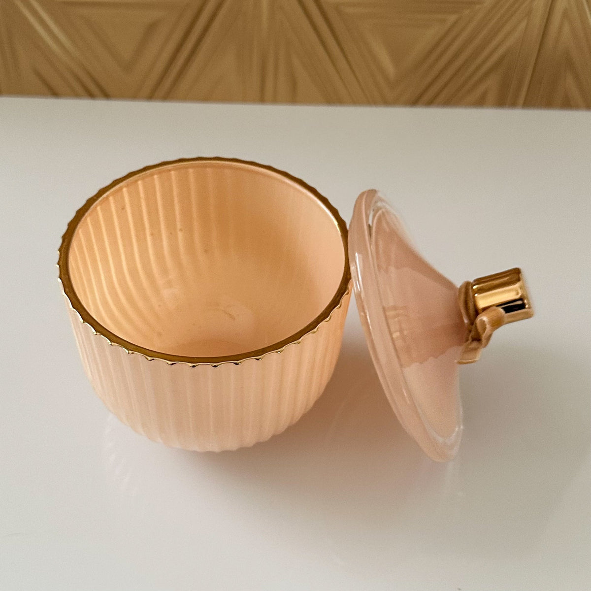 A small ribbed glass container in a pale peach-pink color with a gold rim, shown open with its matching lid leaning against it on a white surface.