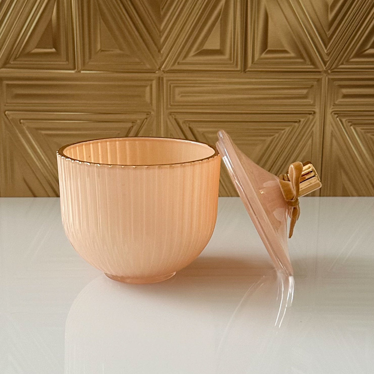 A ribbed peach-colored glass bowl with a matching lid featuring a decorative bow, set on a glossy white surface against a textured gold wall.
