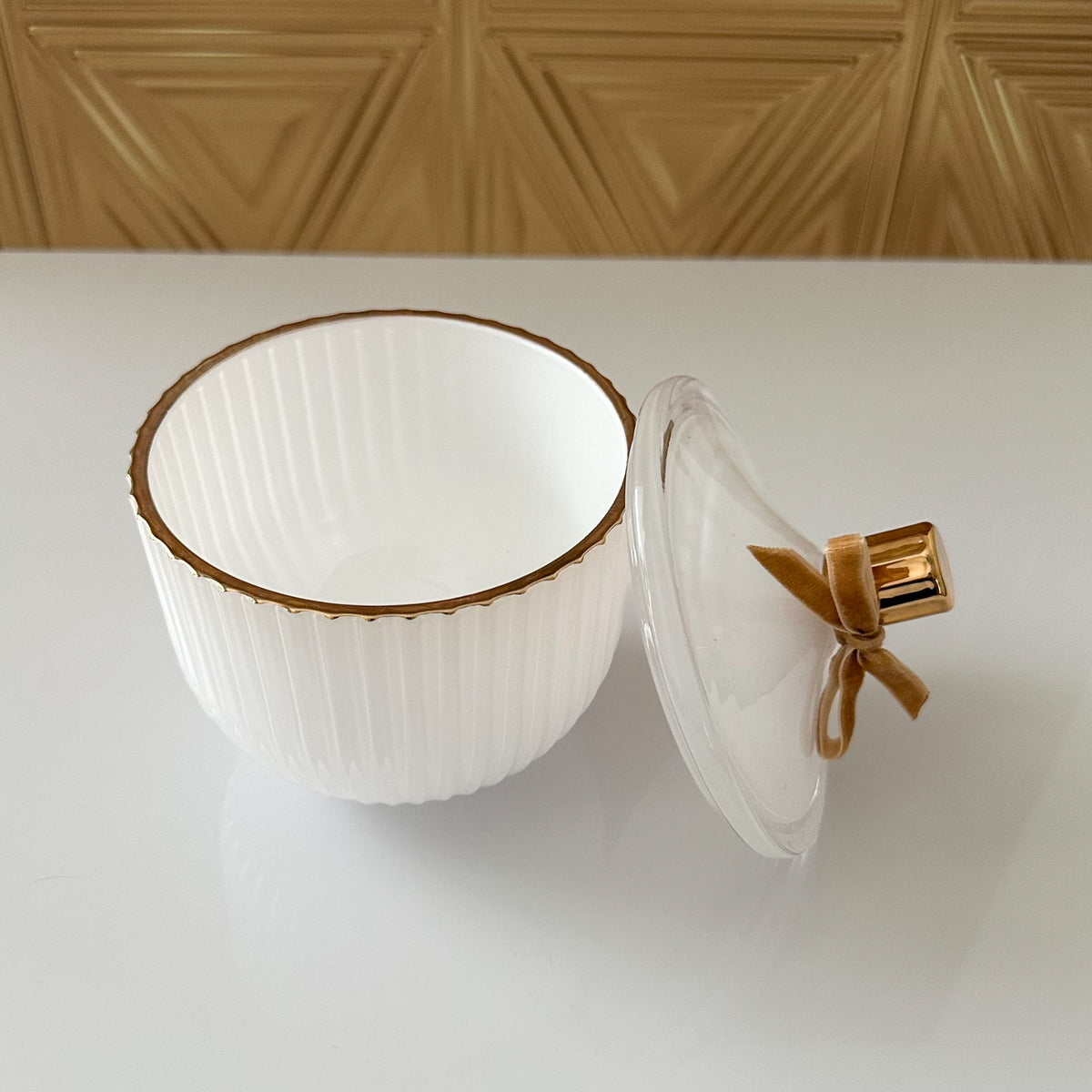 A white ribbed ceramic bowl with a gold rim, shown open with its matching clear glass lid leaning against it on a white surface. The background features a textured gold wall panel.