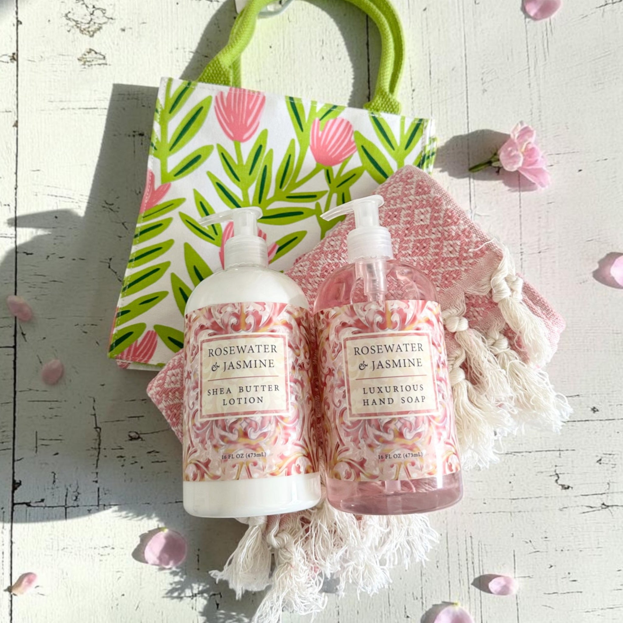 A gift set featuring a white bottle of Rosewater & Jasmine Shea Butter Lotion and a pink bottle of Rosewater & Jasmine Luxurious Hand Soap, styled with a pink and green patterned gift bag, a pink woven pouch with white tassels, and scattered pink flower petals on a white wooden surface.