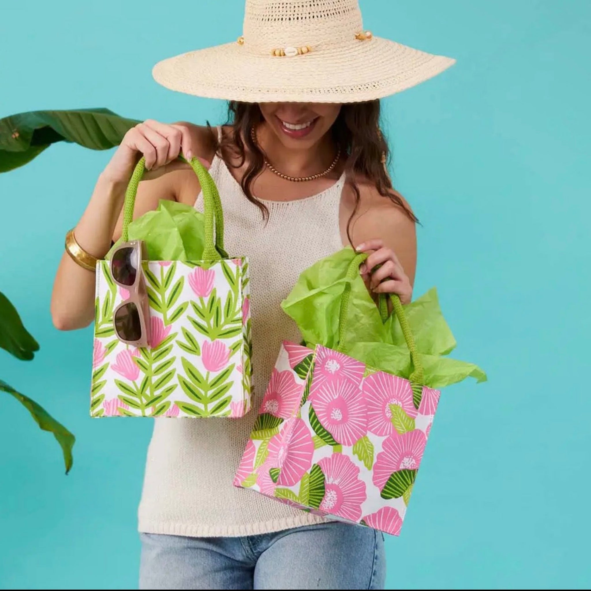A woman wearing a wide-brim straw hat holds two small tote bags with pink tropical floral prints and green leaf patterns. The bags are filled with bright green tissue paper, and the background is a solid turquoise blue with a leafy plant on the left.