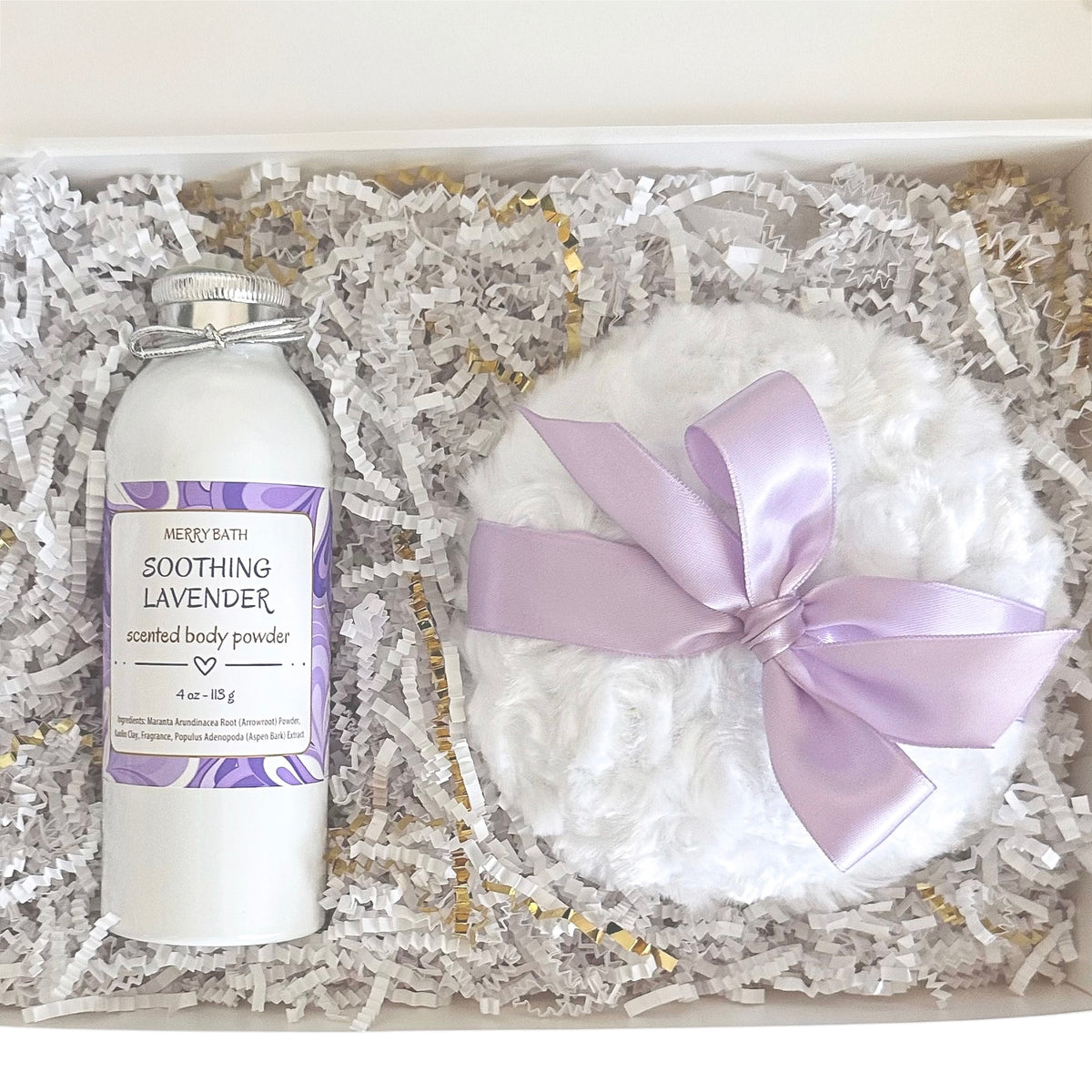 A gift set in a white box filled with white shredded paper, featuring a white bottle of Merry Bath Soothing Lavender scented body powder and a round white body powder puff tied with a light purple satin bow.