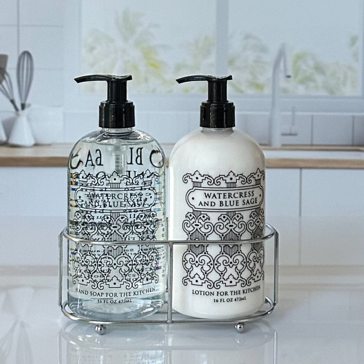Two decorative pump bottles of bath and body products in a clear wire rack on a white countertop, with a blurred kitchen sink background. The left bottle is clear with black ornamental text and liquid inside, and the right bottle is white with matching black text and cream-colored lotion inside.
