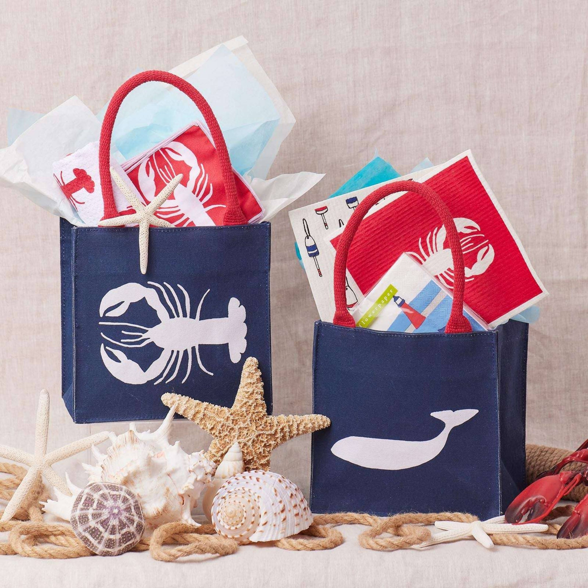 Two navy blue bags with lobster and whale designs, filled with items, surrounded by starfish and rope on a neutral background.