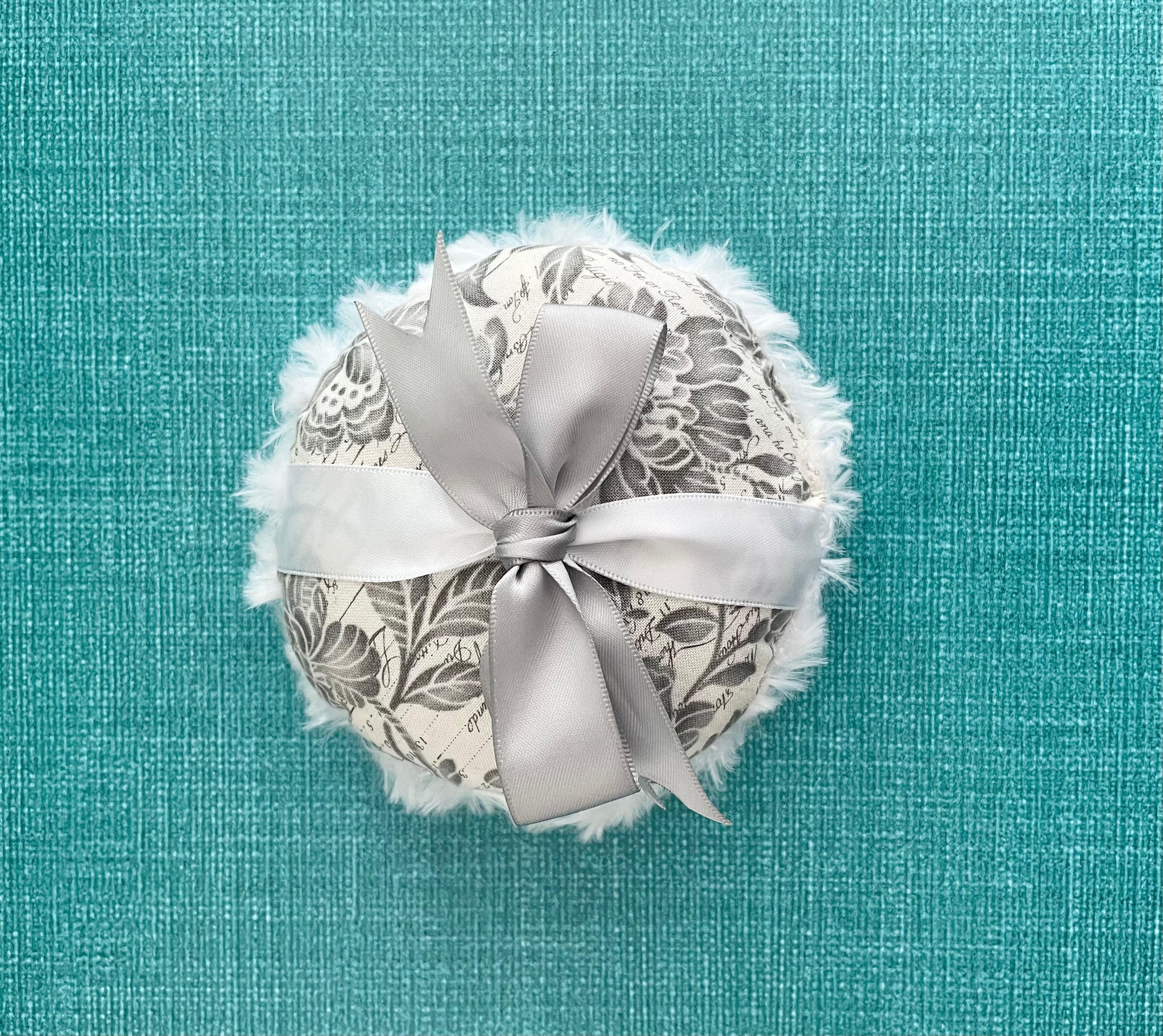 Round powder puff with gray floral pattern, white lace trim, and center satin ribbon bow